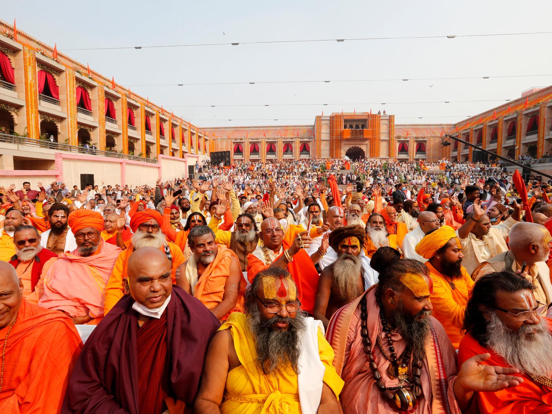Hindu-Männer in langen Gewändern in Gelb-, Orange- und Rottönen sitzen dicht gedrängt auf einem großen Hof.