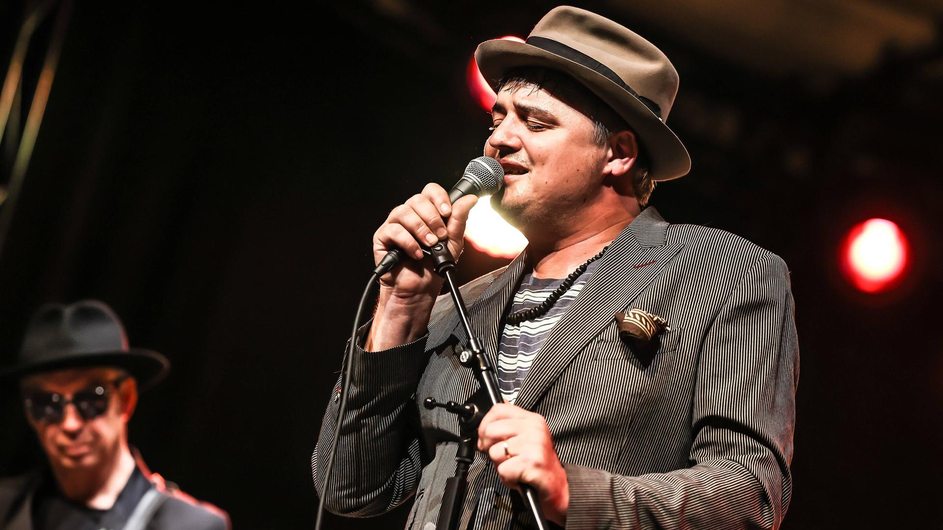 Ein Mann im grauen Anzug und Hut steht auf einer Bühne und singt mit geschlossenen Augen. Es ist der Brite Peter Doherty. Ein Mann im grauen Anzug und Hut steht auf einer Bühne und singt mit geschlossenen Augen. Es ist der Brite Peter Doherty.