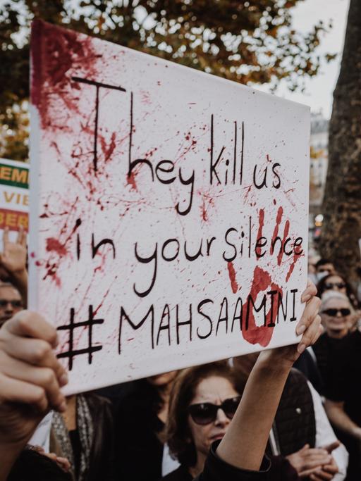 Demonstrierende halten Plakate mit der Aufschrift "They kill us in your silence" und "Femme Vie, Lieberté"