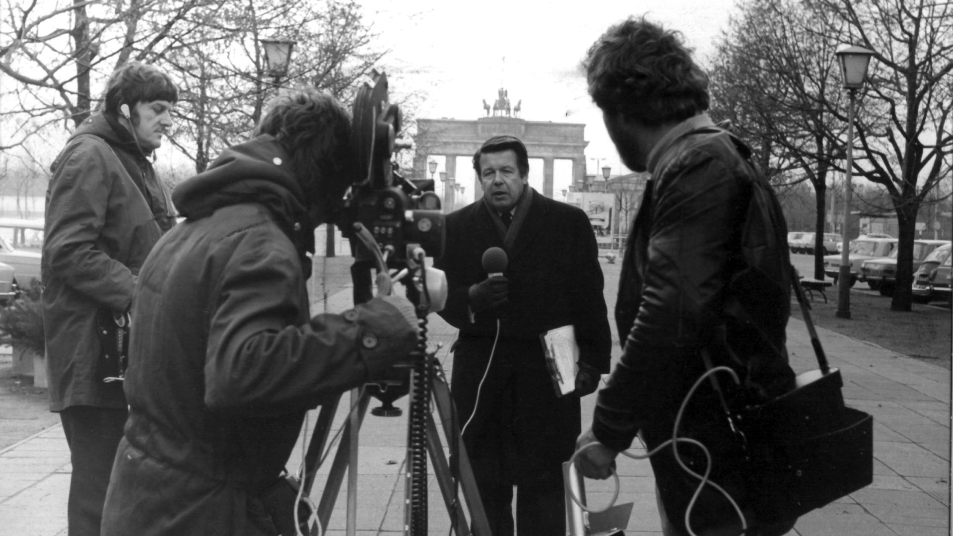 Der Journalist Lothar. Ein Kamerateam und ein Reporter mit Mikrofon in der Hand stehen vor dem Brandenburger Tor. Der Journalist Lothar. Ein Kamerateam und ein Reporter mit Mikrofon in der Hand stehen vor dem Brandenburger Tor.