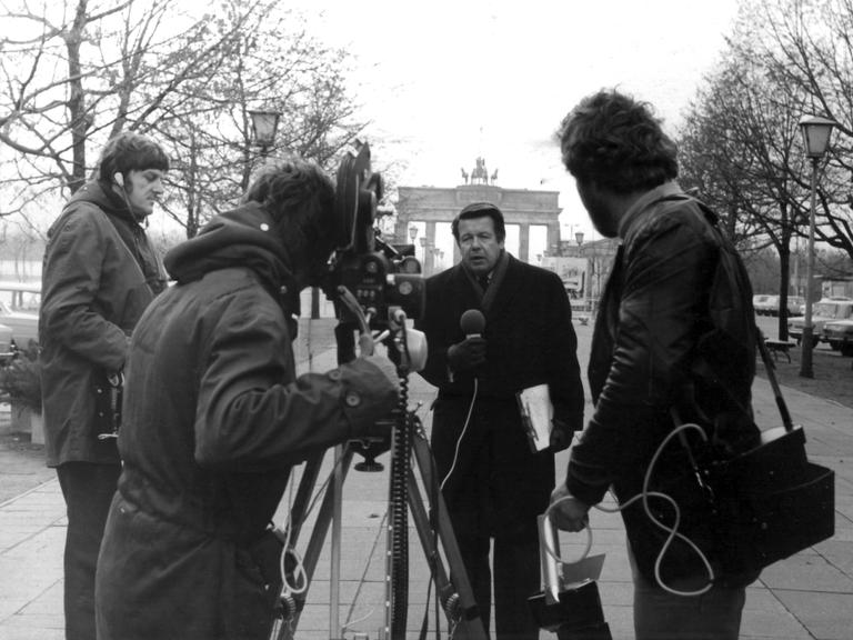 Der Journalist Lothar. Ein Kamerateam und ein Reporter mit Mikrofon in der Hand stehen vor dem Brandenburger Tor.
