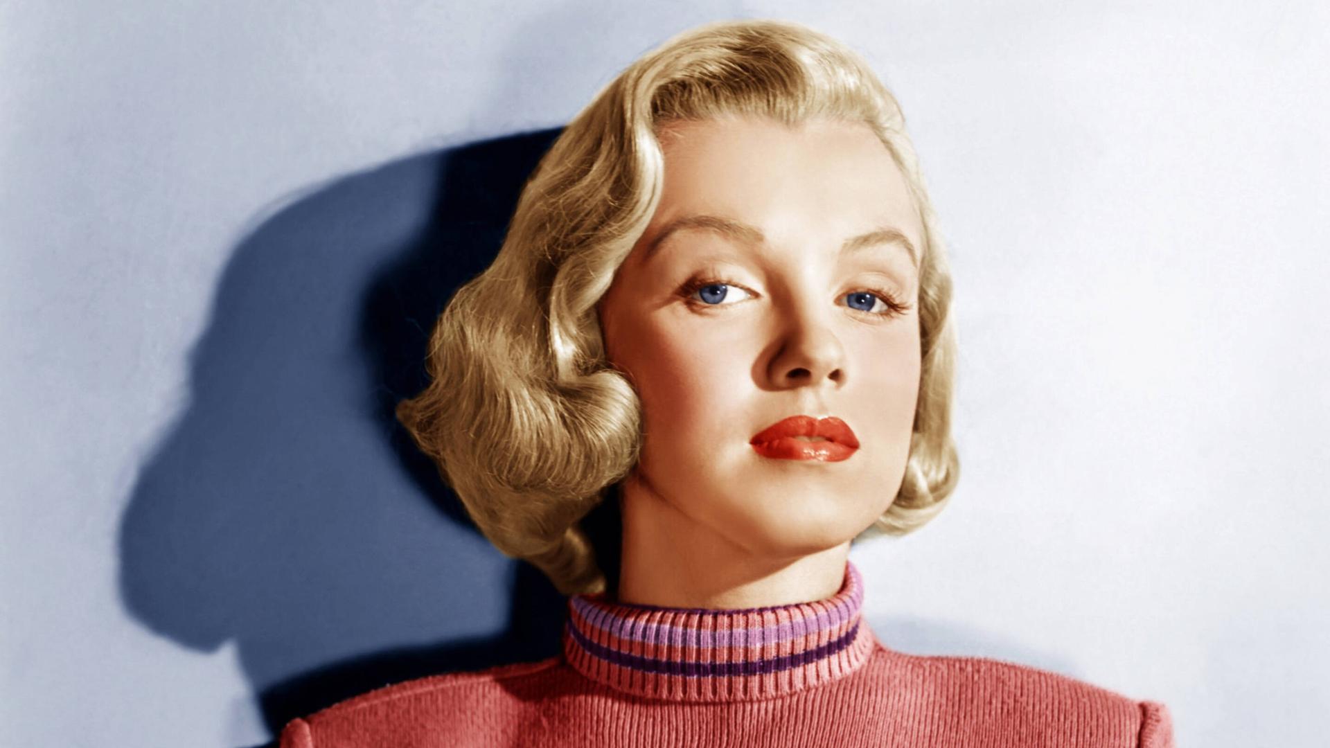 Marilyn Monroe steht in einem rosafarbenen Pullover an eine Wand gelehnt, ihr Haar ist blond gefärbt. 