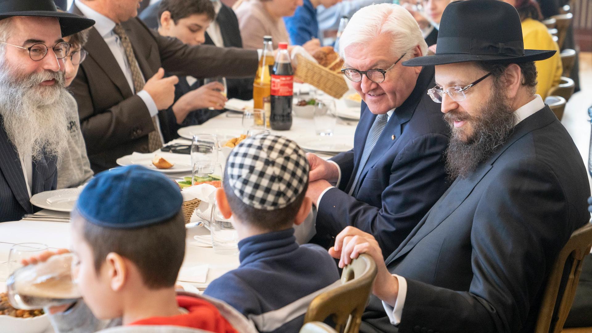 Frank-Walter Steinmeier sitzt mit orthodox gekleideten Juden an einem Tisch beim Essen und spricht mit aus der Ukraine geflüchteten Kindern. Frank-Walter Steinmeier sitzt mit orthodox gekleideten Juden an einem Tisch beim Essen und spricht mit aus der Ukraine geflüchteten Kindern.
