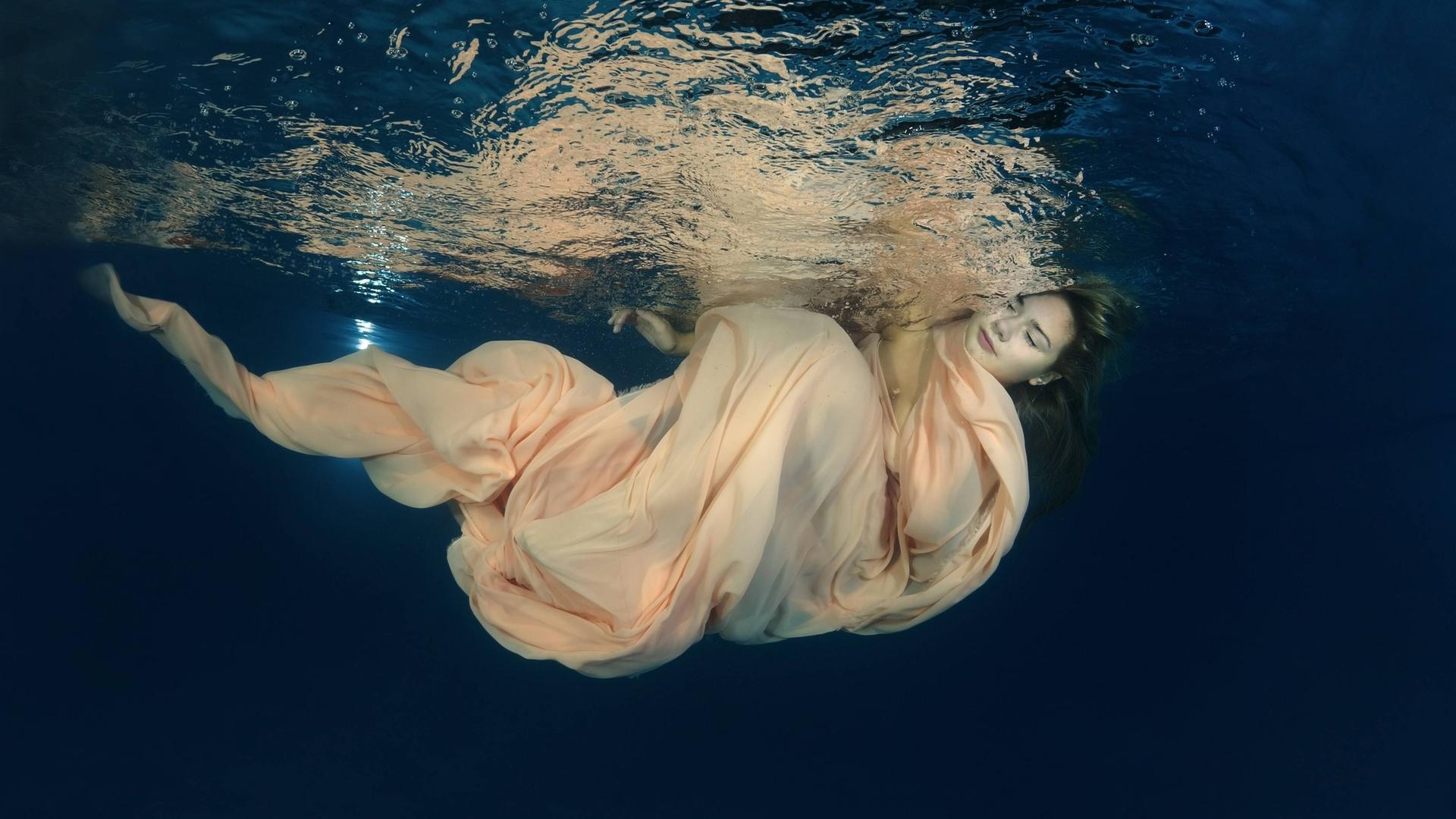 Eine Frau in einem wallenden Kleid unter der Wasseroberfläche, ihre Augen sind geschlossen