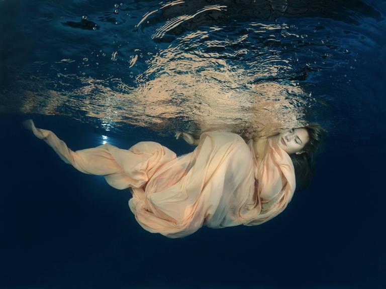 Eine Frau in einem wallenden Kleid unter der Wasseroberfläche, ihre Augen sind geschlossen