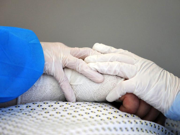 Eine Nahaufnahme der verbundenen Hand eines Covid-19-Patienten, auf der zwei Hände in Schutzhandschuhen sanft aufliegen.