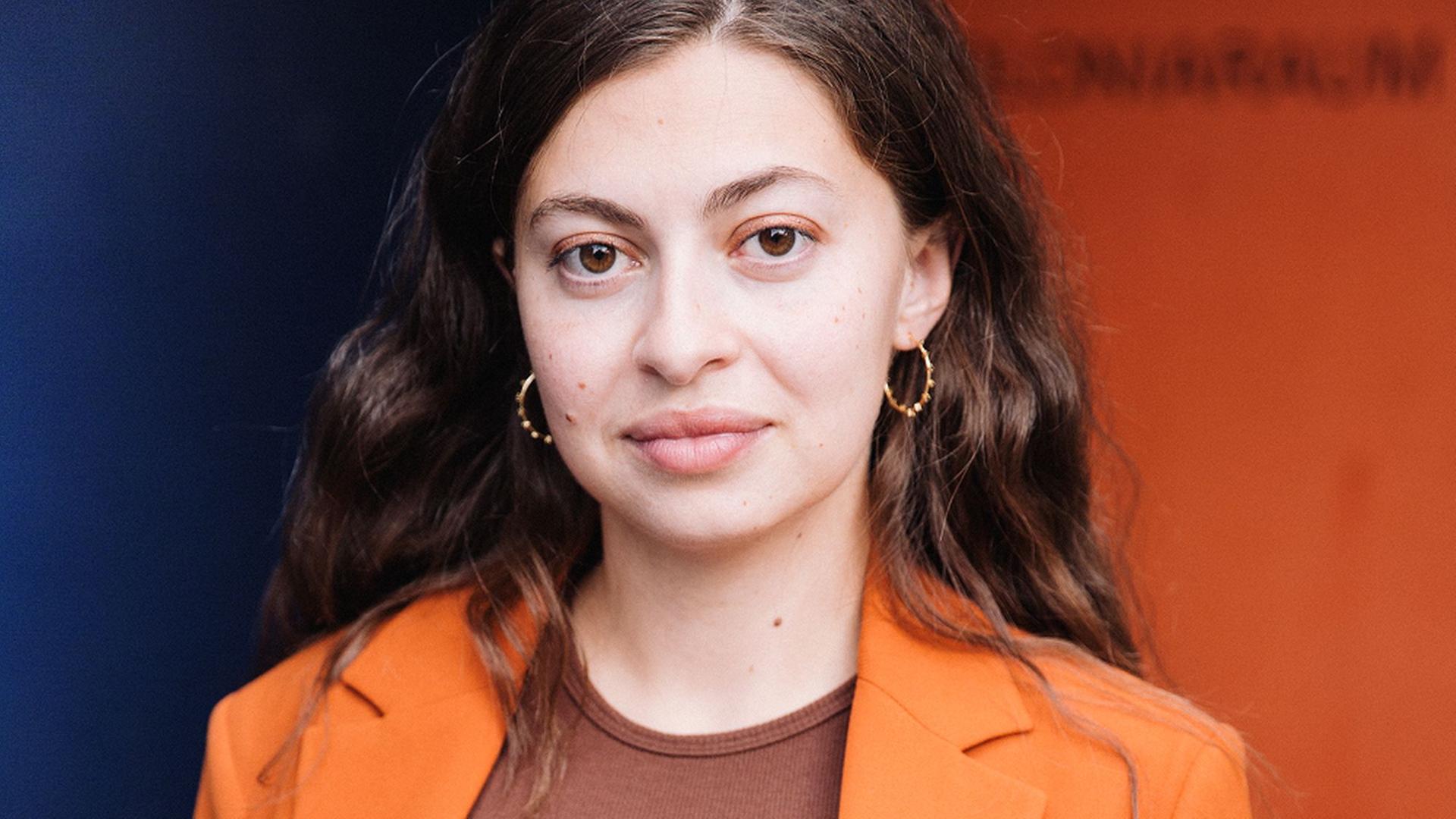 Porträt von Luna Al-Mousli. Eine junge Frau mit glatten, dunkelbraunen Haaren, die einen orangen Blazer trägt. Porträt von Luna Al-Mousli. Eine junge Frau mit glatten, dunkelbraunen Haaren, die einen orangen Blazer trägt.
