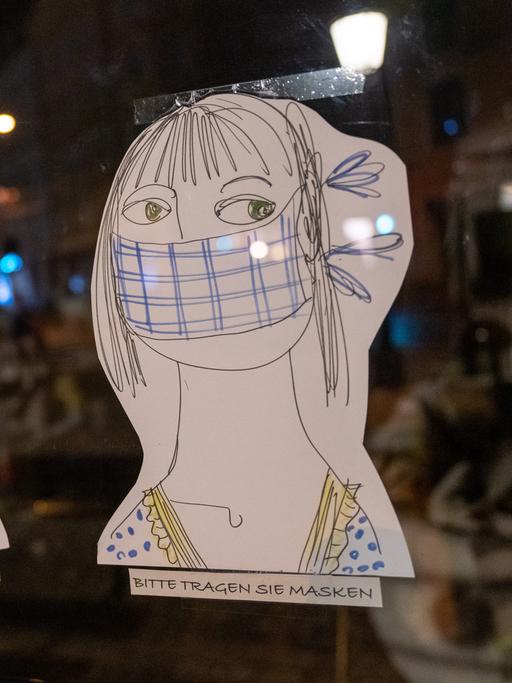 Am Schaufenster eines Geschäfts kleben drei auf Papier gezeichnete und ausgeschnittene Köpfe, darunter jeweils der handschriftliche Hinweis: "Bitte tragen Sie Masken".