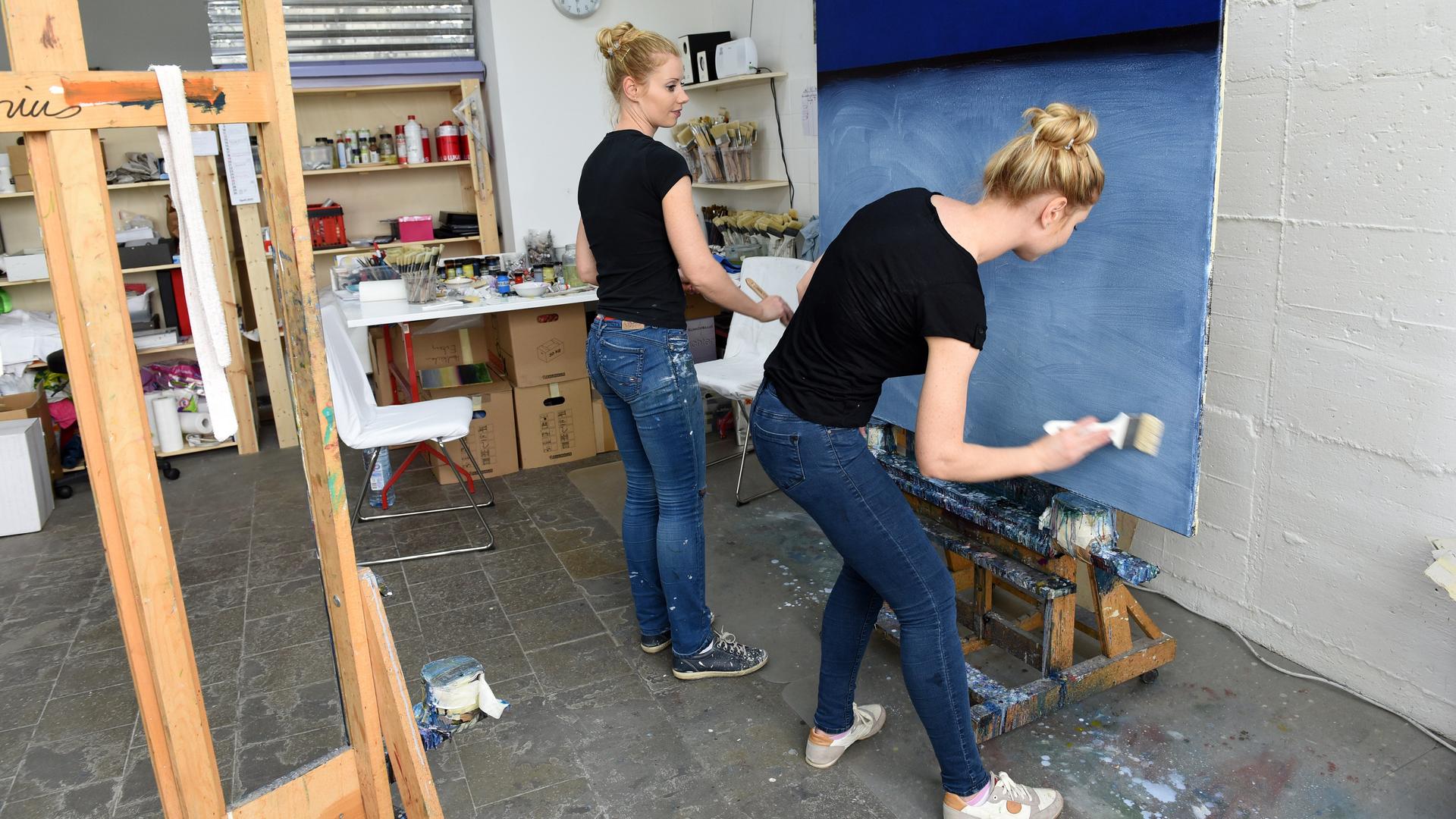 Zwei blonde Frauen in Jeans und schwarzem T-Shirt, Irina (l.) und Marina Fabrizius, malen gemeinsam an einem Bild in Düsseldorf. Das Bild ist in verschiedenen Blautönen gehalten. Links im Vordergrund ist eine Staffelei im Bild.