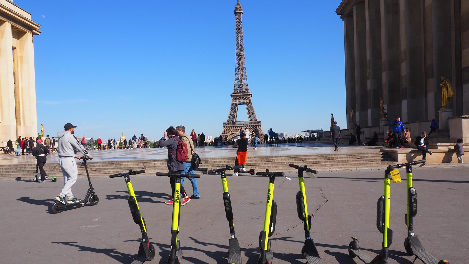 Das Foto zeigt gelbe E-Roller, die vor dem Eiffelturm auf einem Platz stehen. Das Foto zeigt gelbe E-Roller, die vor dem Eiffelturm auf einem Platz stehen.