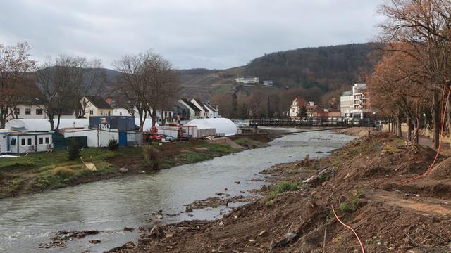 Von der Flut 2021 beschädigte Häuser, Container und weitere Behelfsbauten stehen bei Dernau am Ufer der Ahr, im Vordergrund aufgewühlte Erde mit Schläuchen darauf. 

