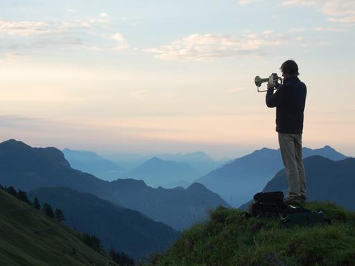 Ein Mann spielt vor einer malerischen Gebirgslandschaft im Dämmerlicht Flügelhorn und ist dabei schräg von hinten zu sehen.
