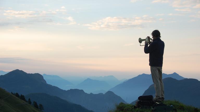 Ein Mann spielt vor einer malerischen Gebirgslandschaft im Dämmerlicht Flügelhorn und ist dabei schräg von hinten zu sehen. Ein Mann spielt vor einer malerischen Gebirgslandschaft im Dämmerlicht Flügelhorn und ist dabei schräg von hinten zu sehen.