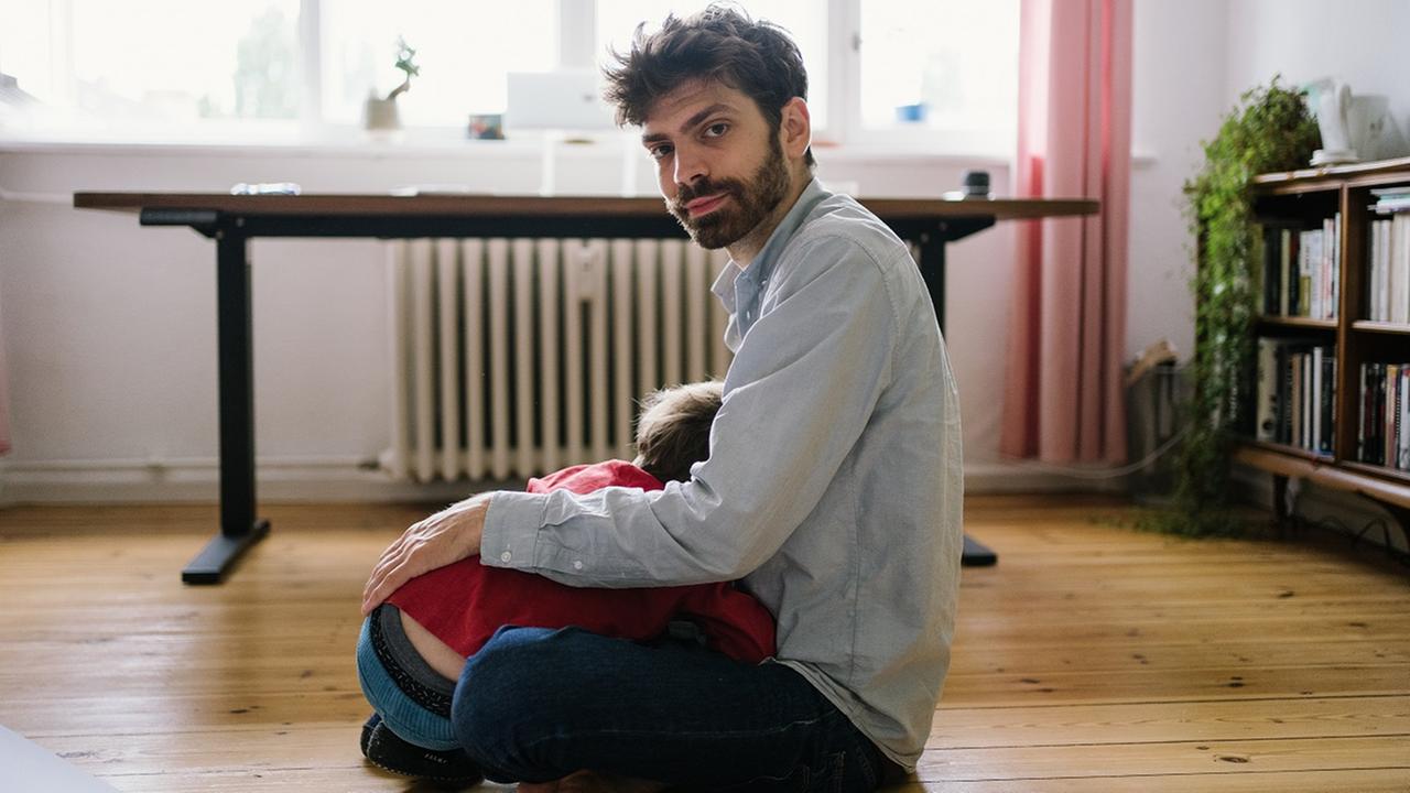 Der Schriftsteller Tobias Wilhelm sitzt mit verschränkten Beinen auf dem Boden, mit abgewandtem Gesicht liegt ein Kind in seinem Schoß, im Hintergrund sind ein Schreibtisch und Gardinen zu sehen. 