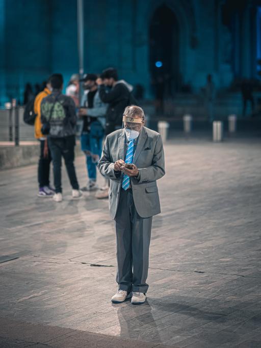 Ein älterer Mann in Anzug und Turnschuhen steht auf einem Platz. Er trägt eine Maske und einen Gesichtsschutz. In der Hand hält er ein Smartphone, auf das er mit gesenktem Kopf guckt.