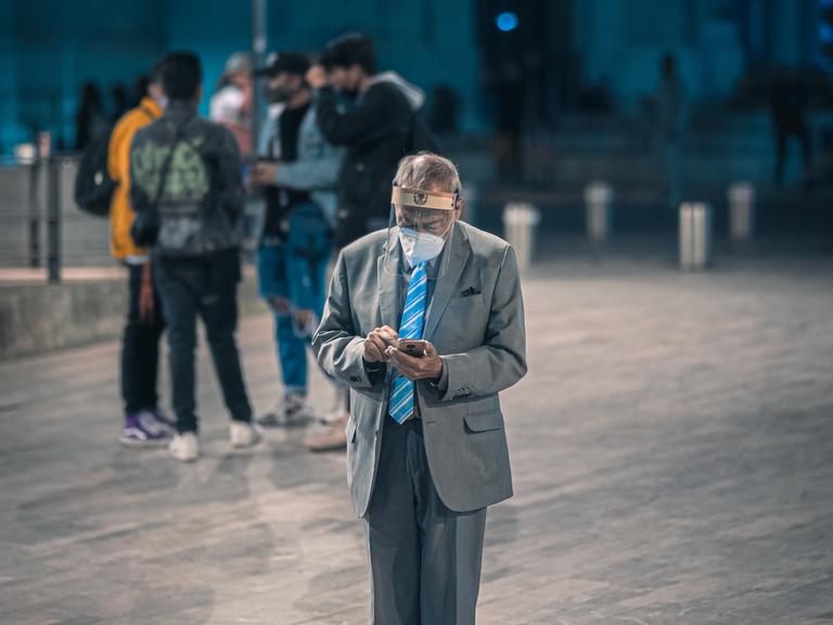 Ein älterer Mann in Anzug und Turnschuhen steht auf einem Platz. Er trägt eine Maske und einen Gesichtsschutz. In der Hand hält er ein Smartphone, auf das er mit gesenktem Kopf guckt.