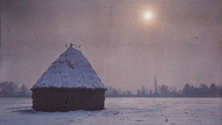 Auf einer frühen Fotografie steht eine Hütte im Schnee vor fahler Sonne.