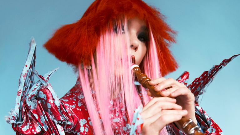 Musikerin Björk spielt ein Blasinstrument. Auf dem Foto hat sie rote und rosa Haare, sie trägt ein kleinteilig gemustertes Gewand, hauptsächlich in der Farbe Rot.