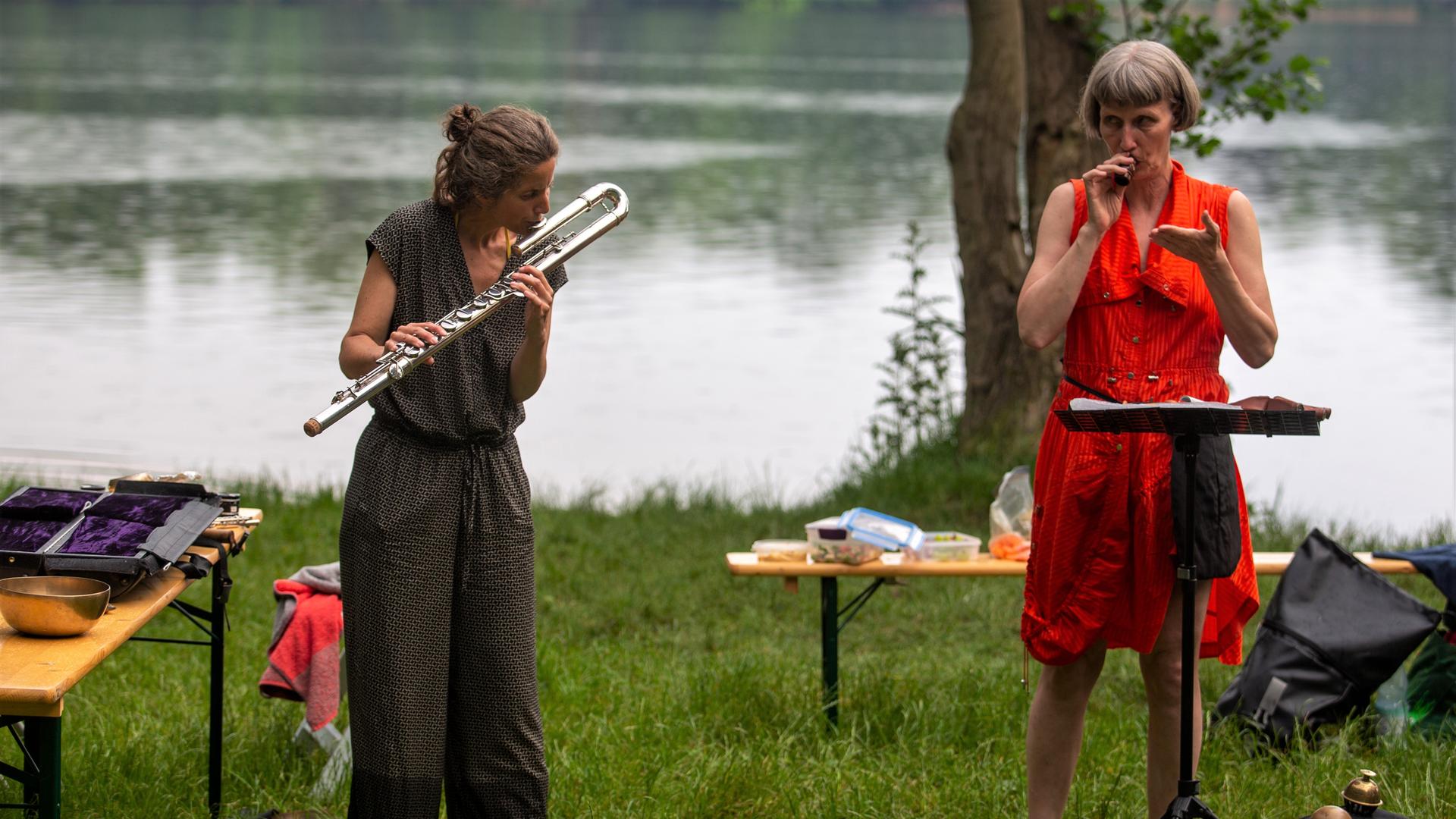 Sabine Vogel und Ute Wassermann stehen inmitten verschiedener Utensilien auf dem Rasen vor dem See und geben ein Konzert Sabine Vogel und Ute Wassermann stehen inmitten verschiedener Utensilien auf dem Rasen vor dem See und geben ein Konzert