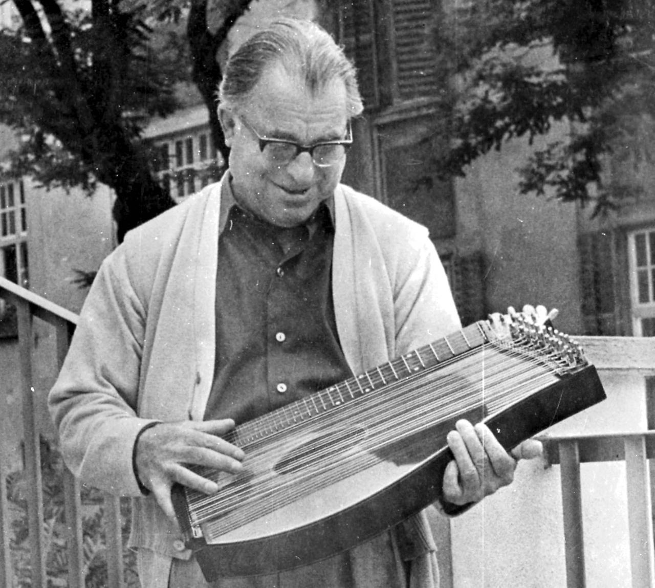 Der ältere Anton Karas steht 1966 mit einer Zither im Arm vor einem Haus mit Treppe und blickt liebevoll auf sein Instrument.