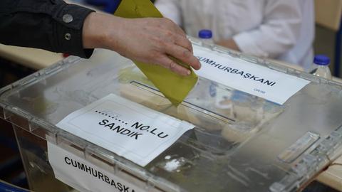 Zu sehen ist eine transparente Wahlbox, in die ein Wahlzettel eingeworfen wird.