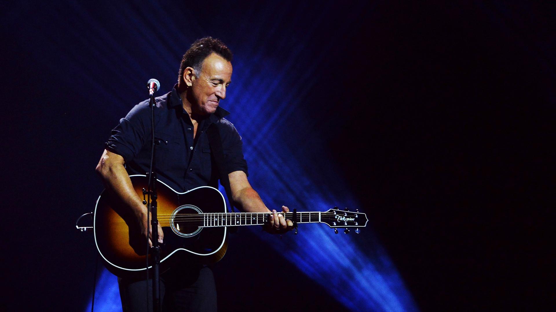 Bruce Springsteen steht auf einer Bühne und spielt eine akustische Gitarre. Der Hintergrund ist in blaues Licht getaucht.
