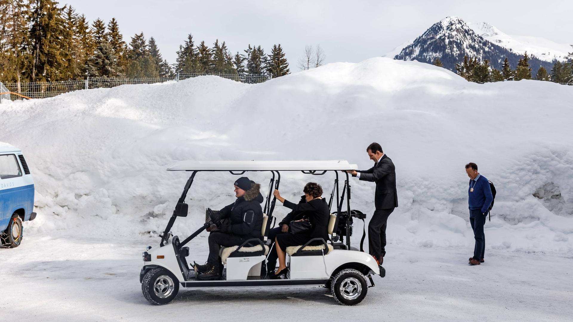 Teuer gekleidete Besucher des Weltwirtschaftsforums in Davos/Schweiz sitzen in einem Golf Trolley und werden durch den Schnee gefahren, im Hintergrund sind Berge zu sehen.