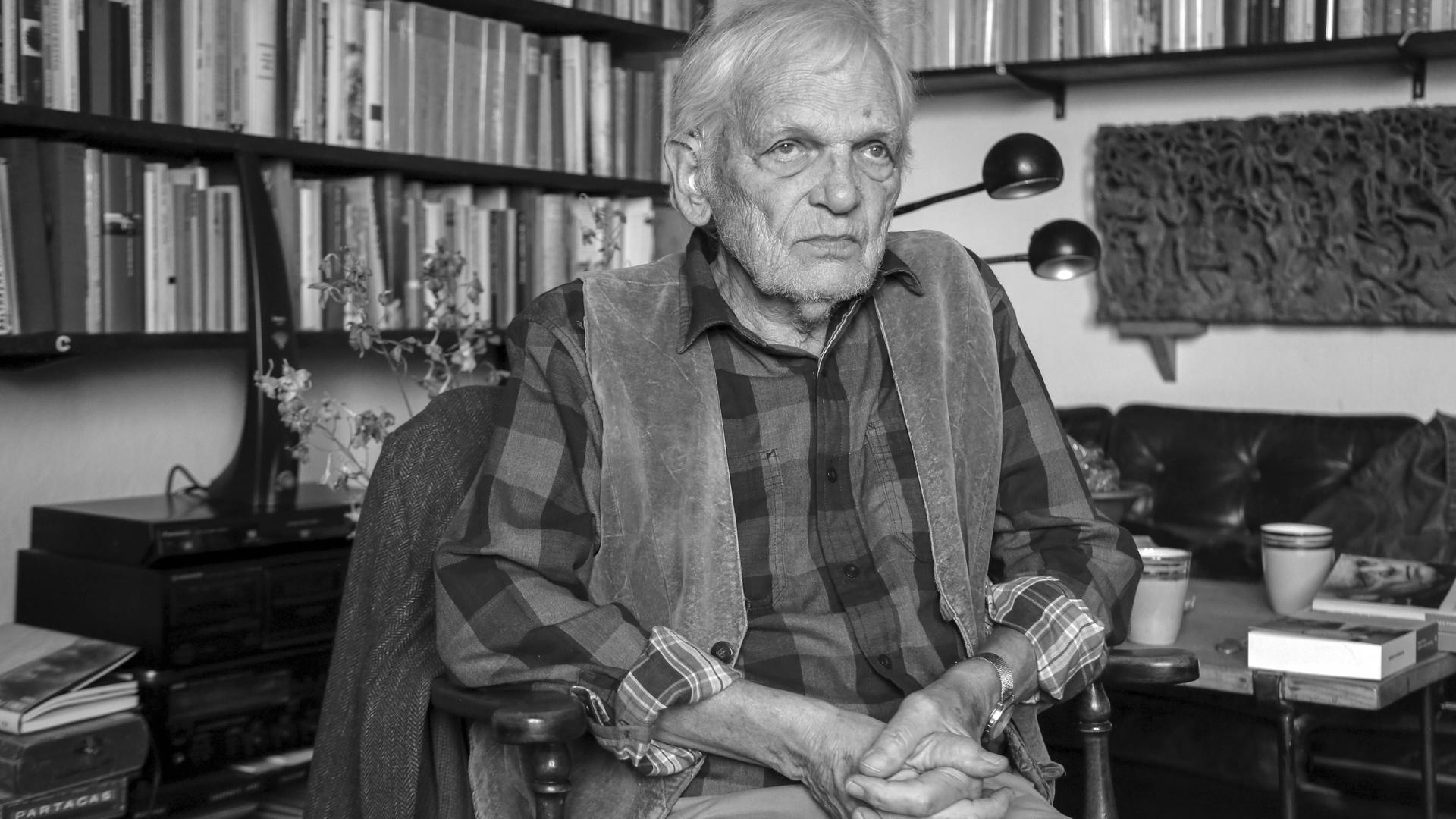 Erasmus Schöfer sitzt in einem Zimmer. Im Hintergrund sind viele Bücher in Regalen zu sehen. Er hat weiße Haare, trägt ein kariertes Hemd und darüber eine Weste. Erasmus Schöfer sitzt in einem Zimmer. Im Hintergrund sind viele Bücher in Regalen zu sehen. Er hat weiße Haare, trägt ein kariertes Hemd und darüber eine Weste.