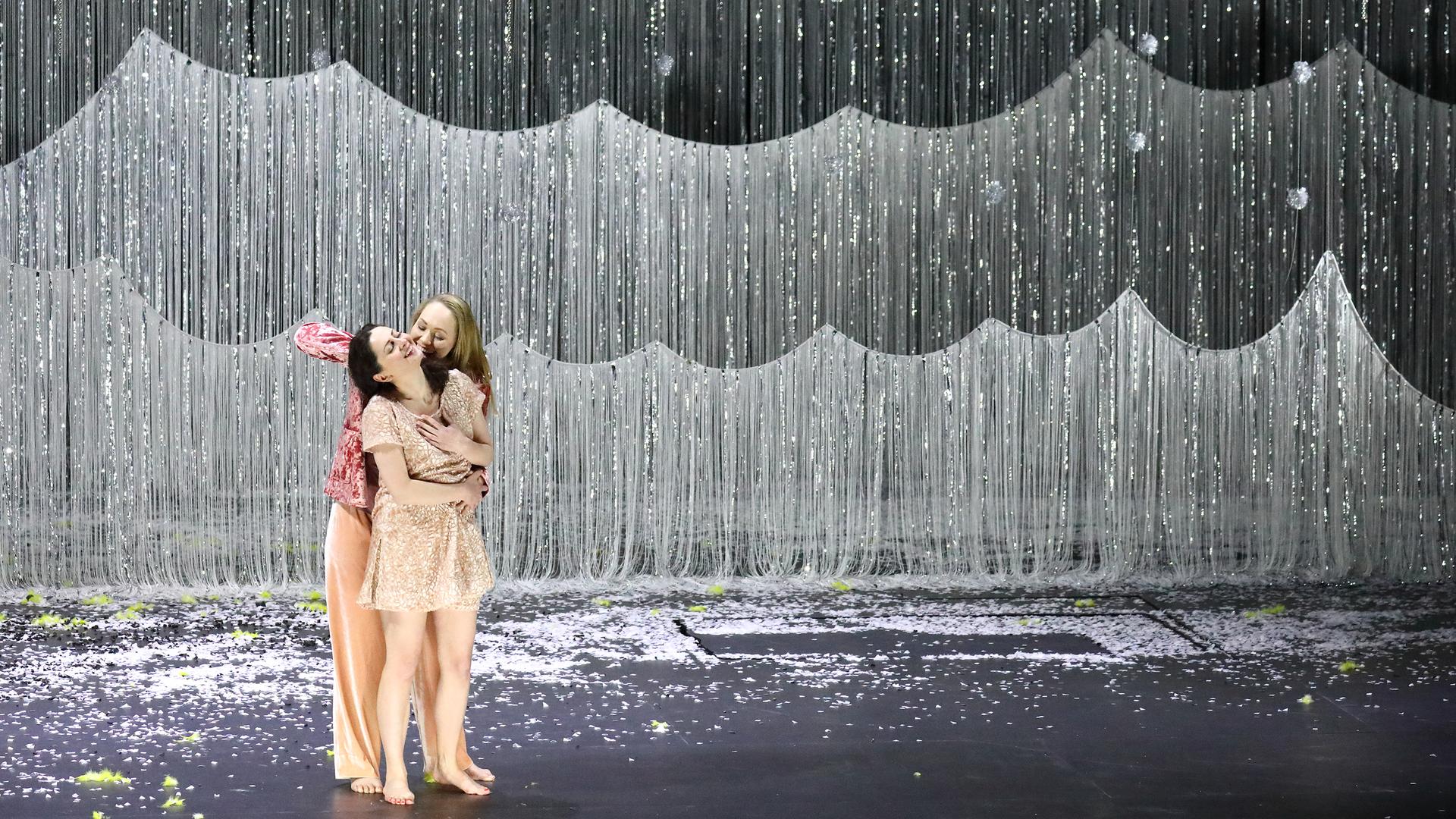 Zwei Frauen umarmen sich auf einer Bühne. Im Hintergrund hängt ein Glitzervorhang als Wald.