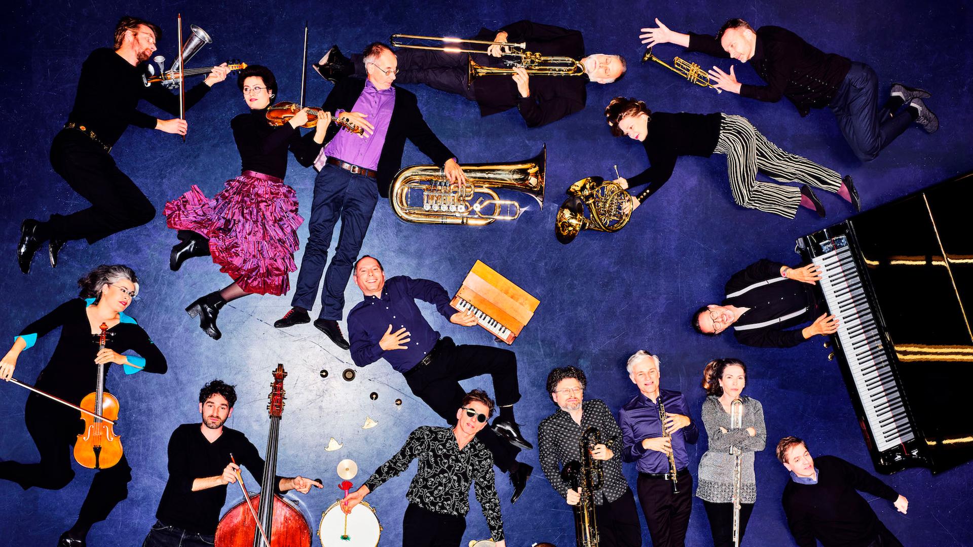 Blick aus der Vogelperspektive auf 15 Musikerinnen und Musiker, die mit ihren Instrumenten auf blauem Boden liegend posieren.
