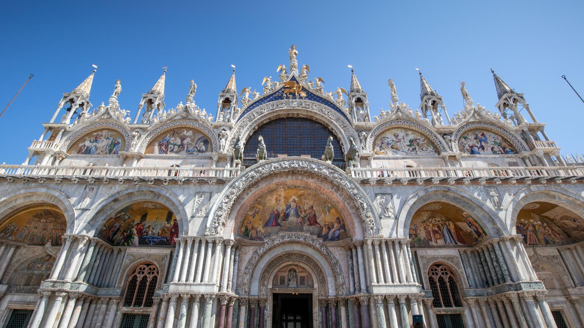 Blick von unten auf die weiße Eingangsfassade des Markusdomes in Venedig, die sich in den blauen Himmel reckt.