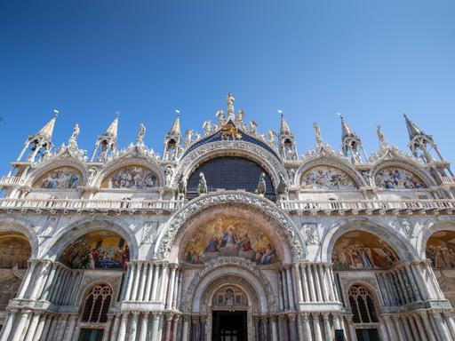 Blick von unten auf die weiße Eingangsfassade des Markusdomes in Venedig, die sich in den blauen Himmel reckt.