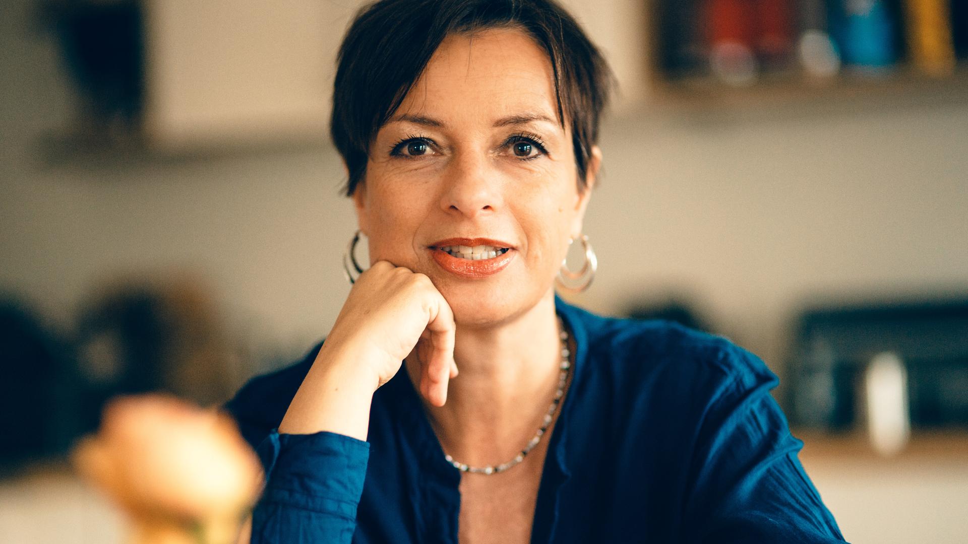 Miriam Gebhardt sitzt an einem Tisch, hat den rechten Ellenbogen auf dem Tisch abgestützt und hält ihr rechte Hand stützend ans Kinn. Sie hat dunkle Haaer, trägt große, rund Ohrringe und schaut in die Kamera. Sie trägt ein blaues Oberteil. Miriam Gebhardt sitzt an einem Tisch, hat den rechten Ellenbogen auf dem Tisch abgestützt und hält ihr rechte Hand stützend ans Kinn. Sie hat dunkle Haaer, trägt große, rund Ohrringe und schaut in die Kamera. Sie trägt ein blaues Oberteil.