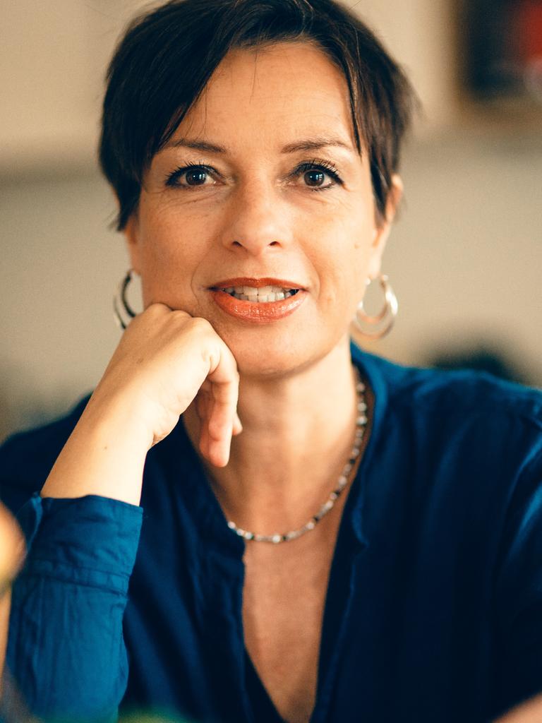 Miriam Gebhardt sitzt an einem Tisch, hat den rechten Ellenbogen auf dem Tisch abgestützt und hält ihr rechte Hand stützend ans Kinn. Sie hat dunkle Haaer, trägt große, rund Ohrringe und schaut in die Kamera. Sie trägt ein blaues Oberteil.