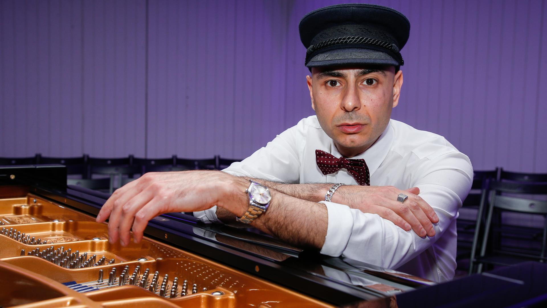 Ein Mann mit einer schwarzen Schiebermütze und weißem Hemd blickt direkt in die Kamera. Er sitzt an einem Flügel, stützt sich auf das Instrument. Ein Mann mit einer schwarzen Schiebermütze und weißem Hemd blickt direkt in die Kamera. Er sitzt an einem Flügel, stützt sich auf das Instrument.