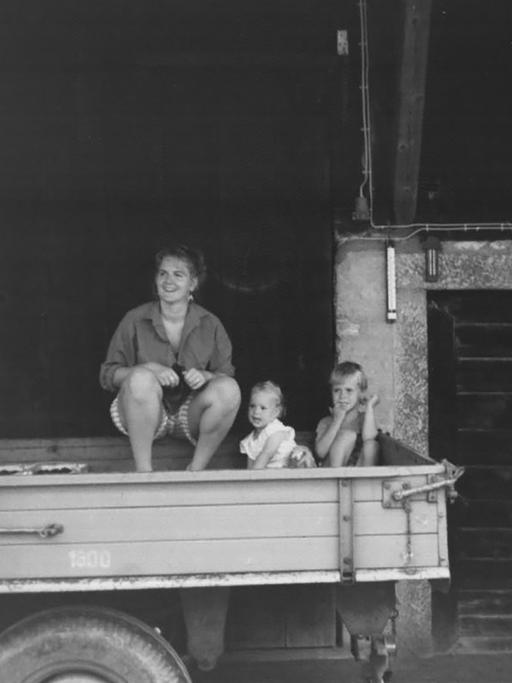 Gabriele sitzt mit zwei Kindern auf einem Hänger, der in einer Scheune steht