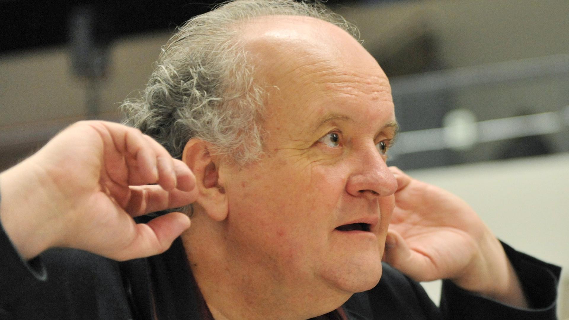 Ein älterer Mann mit schütterem Haar blickt aufmerksam. Er hält seine Hände an die Ohren, um besser zu hören. Es ist der Komponist Wolfgang Rihm. Ein älterer Mann mit schütterem Haar blickt aufmerksam. Er hält seine Hände an die Ohren, um besser zu hören. Es ist der Komponist Wolfgang Rihm.