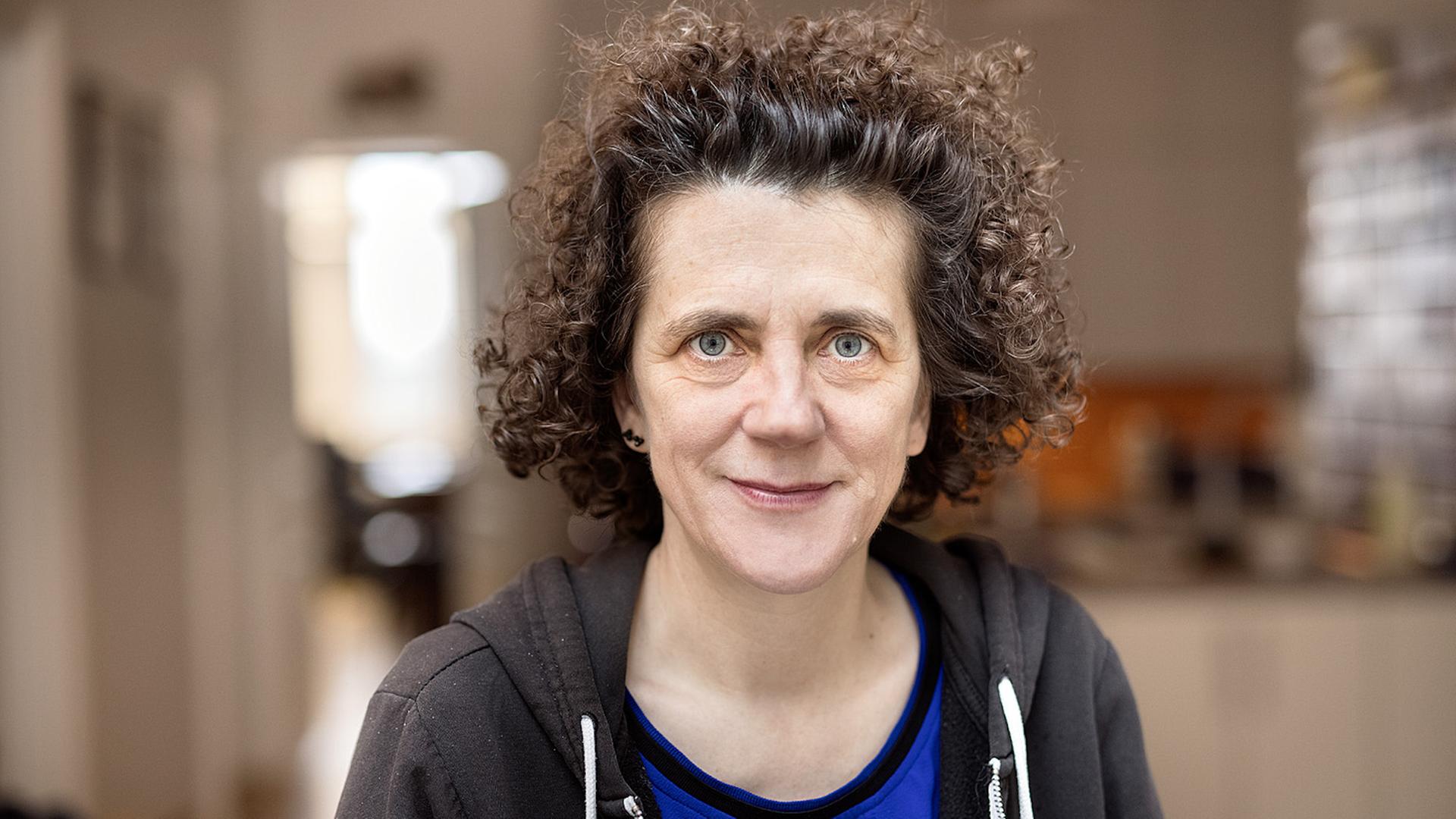 Olga Neuwirth, eine Frau mit lockigen, aus der Stirn nach oben gekämmten Haaren, schaut in die Kamera. Der Hintergrund ist unscharf. Sie trägt ein blauses T-Shirt und eine dunkelgraue Kapuzenjacke. Olga Neuwirth, eine Frau mit lockigen, aus der Stirn nach oben gekämmten Haaren, schaut in die Kamera. Der Hintergrund ist unscharf. Sie trägt ein blauses T-Shirt und eine dunkelgraue Kapuzenjacke.