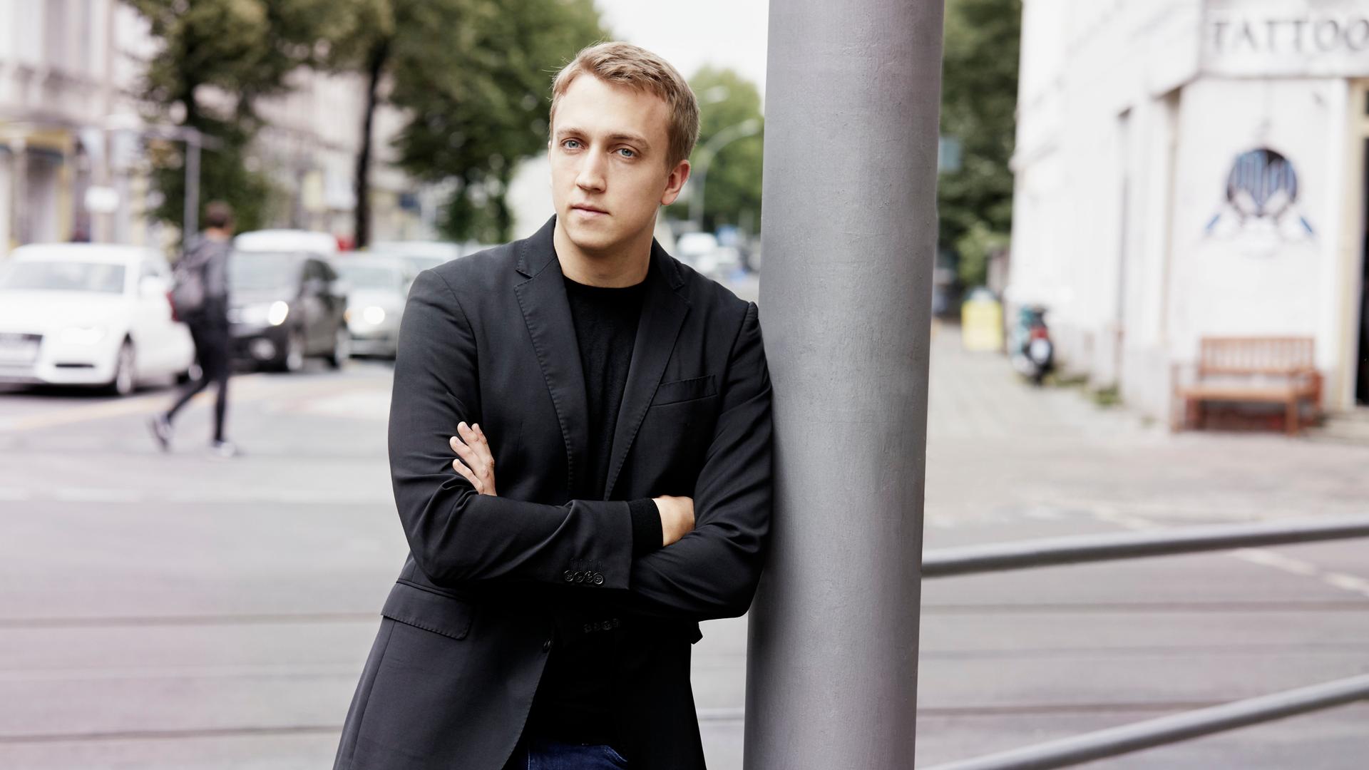Ein junger Mann mit blondem Haar lehnt mit verschränkten Armen an einem silbergrauen Laternenpfahl. Er trägt Jeans und ein schwarzes Jackett. Im Hintergrund ist eine Straße mit geparkten Autos und Bäumen zu sehen. Ein junger Mann mit blondem Haar lehnt mit verschränkten Armen an einem silbergrauen Laternenpfahl. Er trägt Jeans und ein schwarzes Jackett. Im Hintergrund ist eine Straße mit geparkten Autos und Bäumen zu sehen.