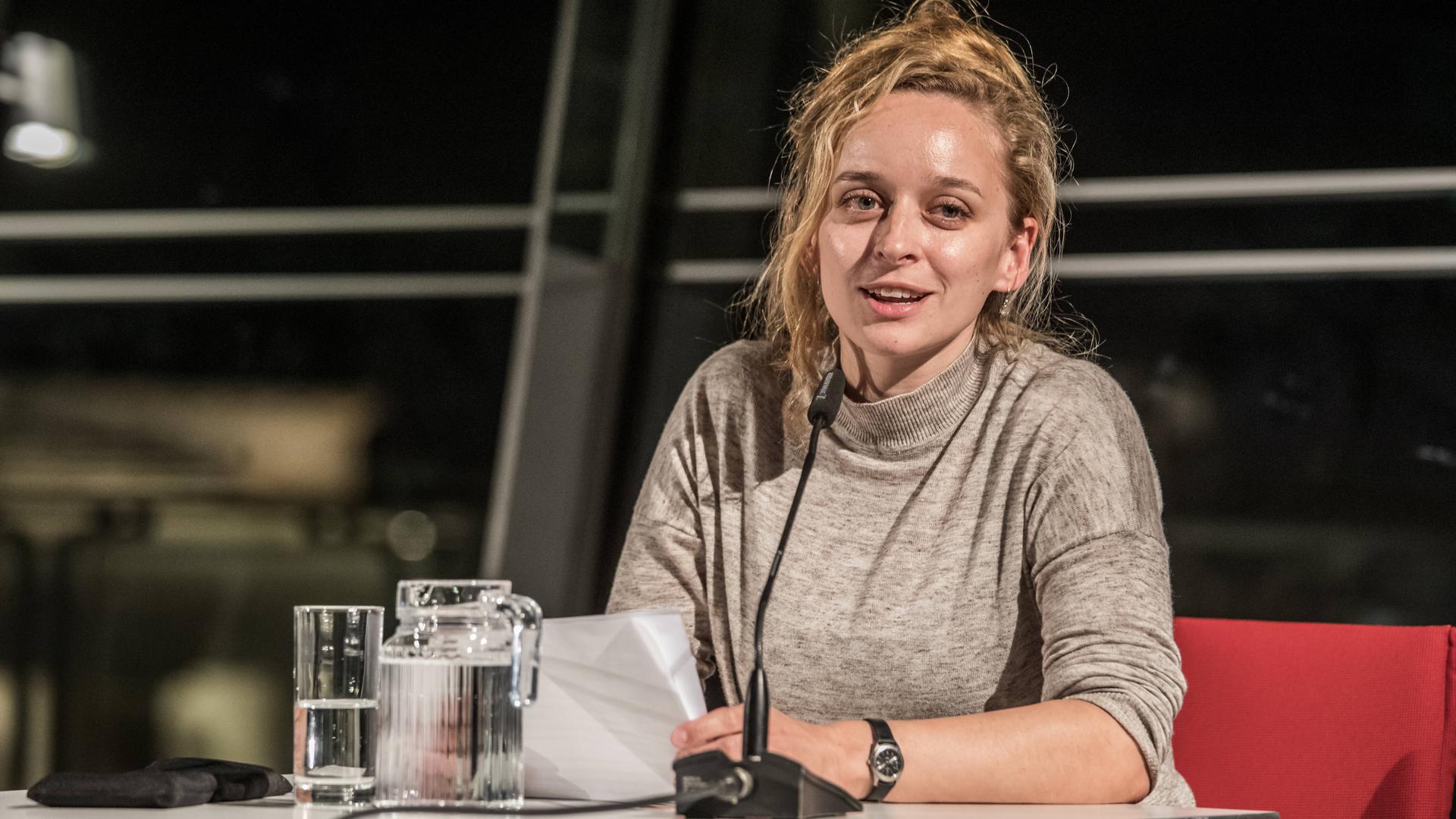 Alina Herbing sitzt in der Berliner Akademie der Künste, vor ihr ein Mikro und Gläser, im Hintergrund eine Fensterfront bei Nacht. Sie trägt einen Pullover in hellen Farben und hat die Haare hochgesteckt, eine Strähne fällt ihr ins Gesicht. Alina Herbing sitzt in der Berliner Akademie der Künste, vor ihr ein Mikro und Gläser, im Hintergrund eine Fensterfront bei Nacht. Sie trägt einen Pullover in hellen Farben und hat die Haare hochgesteckt, eine Strähne fällt ihr ins Gesicht.