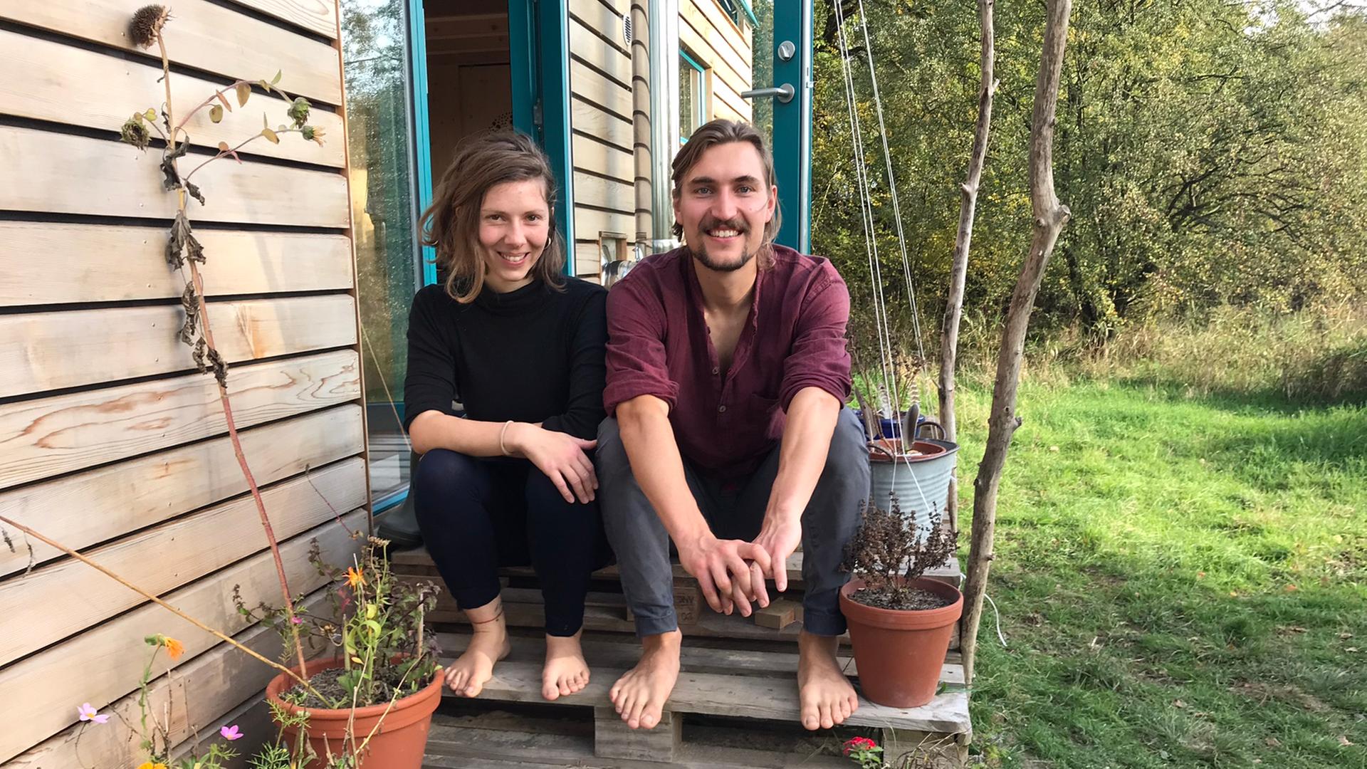 Eine junge Frau und ein junger Mann sitzen auf gestapelten Europaletten, die den Eingang zu ihrem Tiny House bilden, dessen längere Seite links von ihnen zu sehen ist. An den Rändern des Aufgangs stehen Blumentöpfe mit Blumen. Beide sind barfuß, die Frau trägt schwarze Kleidung, der Mann eine grau Hose und ein weinrotes Oberteil. Beide strahlen. Eine junge Frau und ein junger Mann sitzen auf gestapelten Europaletten, die den Eingang zu ihrem Tiny House bilden, dessen längere Seite links von ihnen zu sehen ist. An den Rändern des Aufgangs stehen Blumentöpfe mit Blumen. Beide sind barfuß, die Frau trägt schwarze Kleidung, der Mann eine grau Hose und ein weinrotes Oberteil. Beide strahlen.