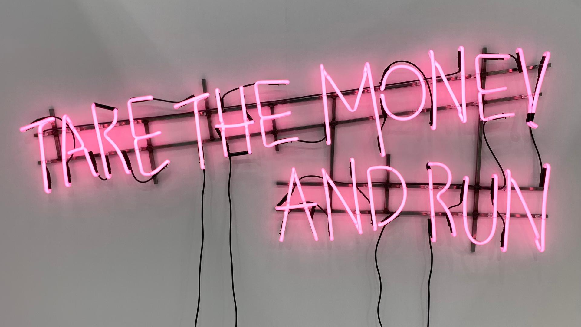 Das Werk „Ne travallez jemais“ (2022) von Kerim Seiler am Stand der Galerie Sexauer auf der Art Cologne. In rosa Buchstaben steht in mit Leuchtstoffröhren vor weisser Wand geschrieben "Take the money and run".