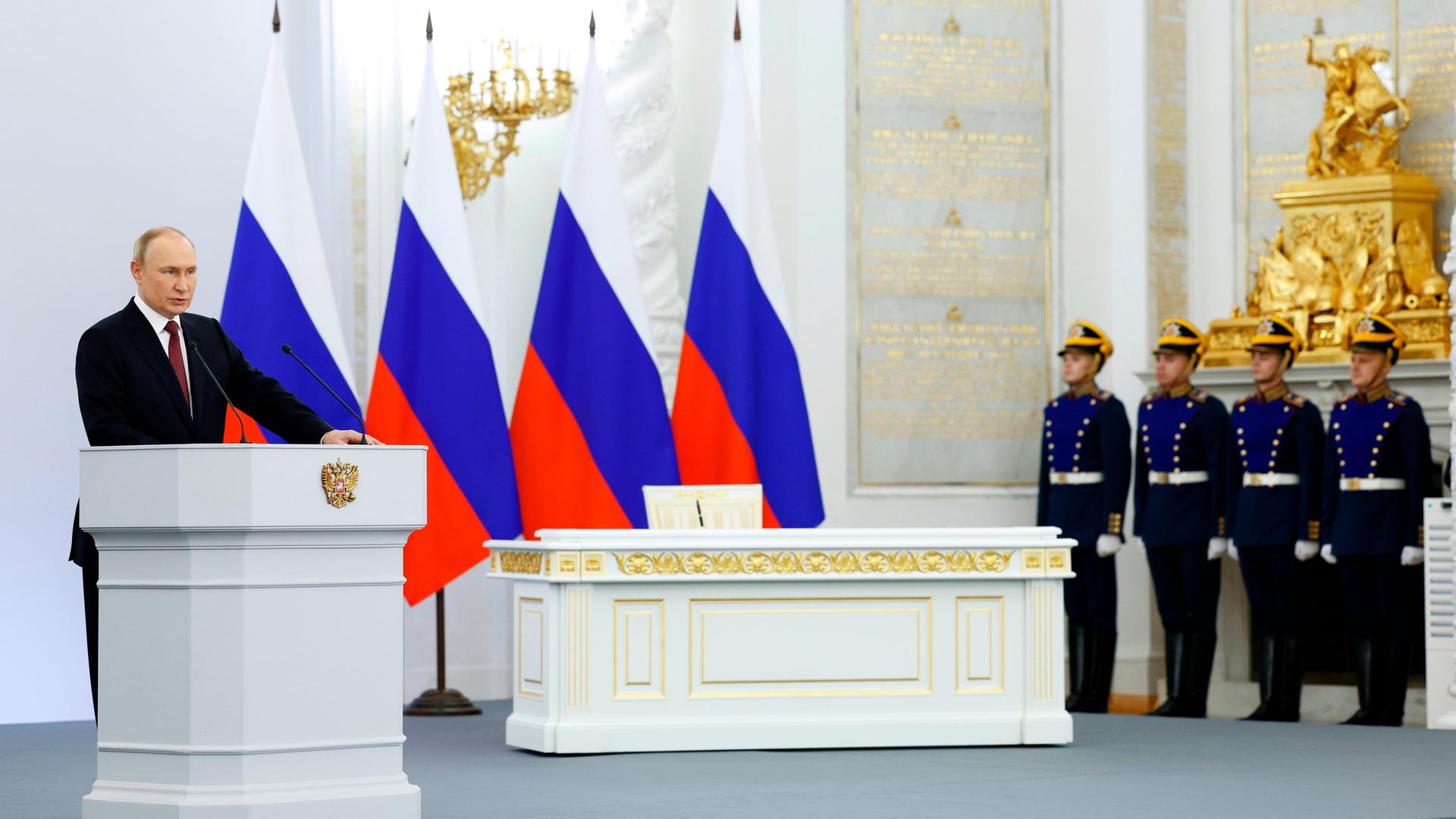 Wladimir Putin steht an einem Rednerpult. Hinter ihm vier russische Flaggen. Neben ihm stehen vier Soldaten. Wladimir Putin steht an einem Rednerpult. Hinter ihm vier russische Flaggen. Neben ihm stehen vier Soldaten.