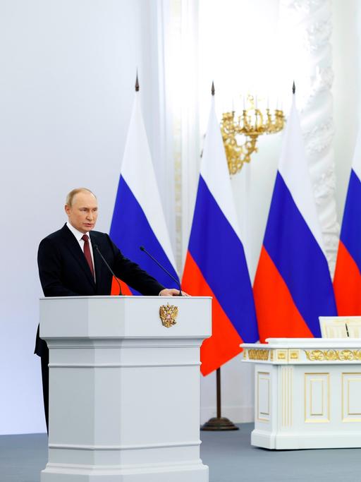 Wladimir Putin steht an einem Rednerpult. Hinter ihm vier russische Flaggen. Neben ihm stehen vier Soldaten.