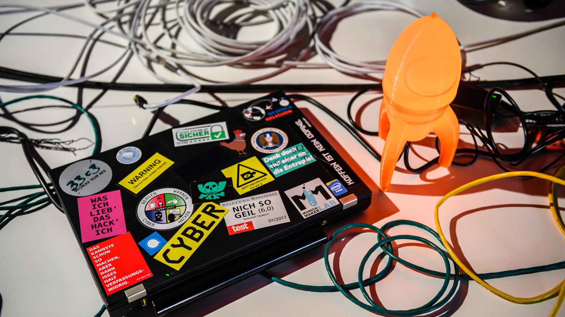 Auf einem Tisch liegt ein Computer der mit vielen Stickern beklebt ist. Auf dem Tisch liegen Kabel und das Maskottchn des CCC - eine Rakete. Auf einem Tisch liegt ein Computer der mit vielen Stickern beklebt ist. Auf dem Tisch liegen Kabel und das Maskottchn des CCC - eine Rakete.
