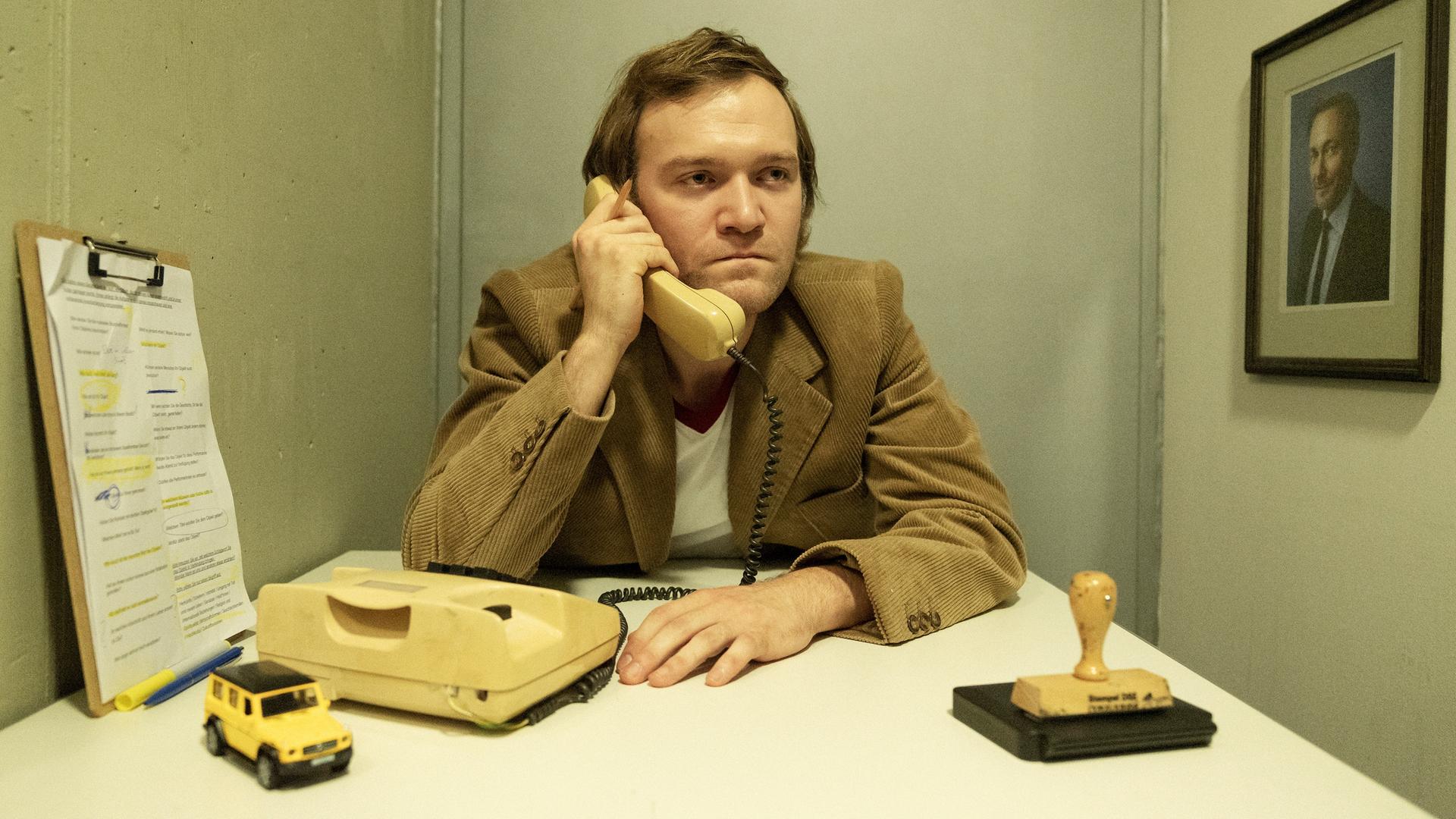 Ein Mann im 70er-Jahre Anzug sitzt an einem Tisch und hält einen Telefonhörer in der Hand. Er sieht nicht sehr glücklich aus. Vor ihm liegt ein Stempelkissen und ein Spielzeugjeep. Ein Mann im 70er-Jahre Anzug sitzt an einem Tisch und hält einen Telefonhörer in der Hand. Er sieht nicht sehr glücklich aus. Vor ihm liegt ein Stempelkissen und ein Spielzeugjeep.