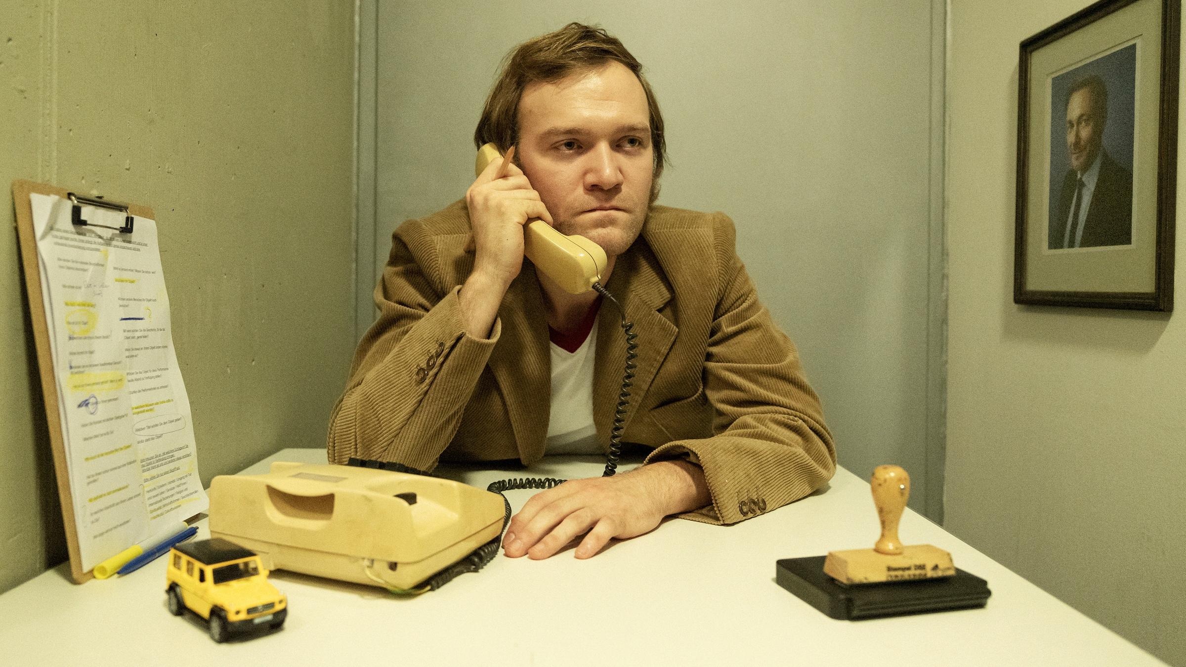 Ein Mann im 70er-Jahre Anzug sitzt an einem Tisch und hält einen Telefo...</p>

                        <a href=