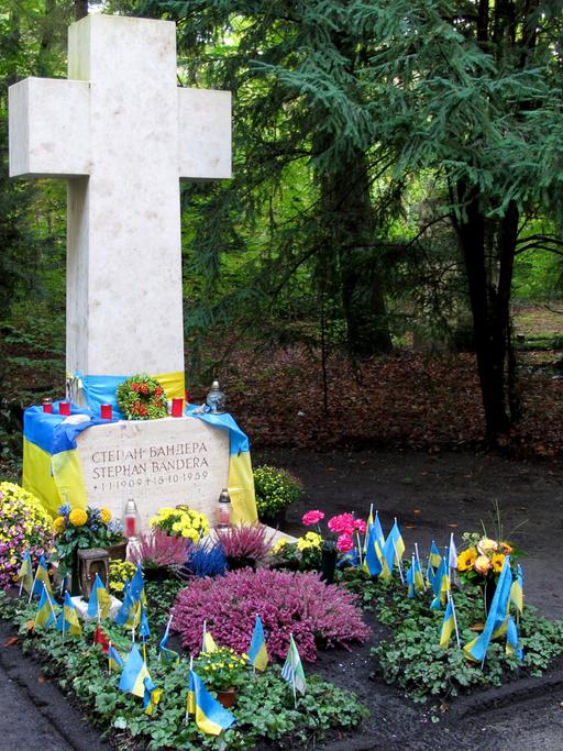 Steinernes Kreuz: Darunter ein mit Blumen und blaugelben Fahnen geschmücktes Grab.