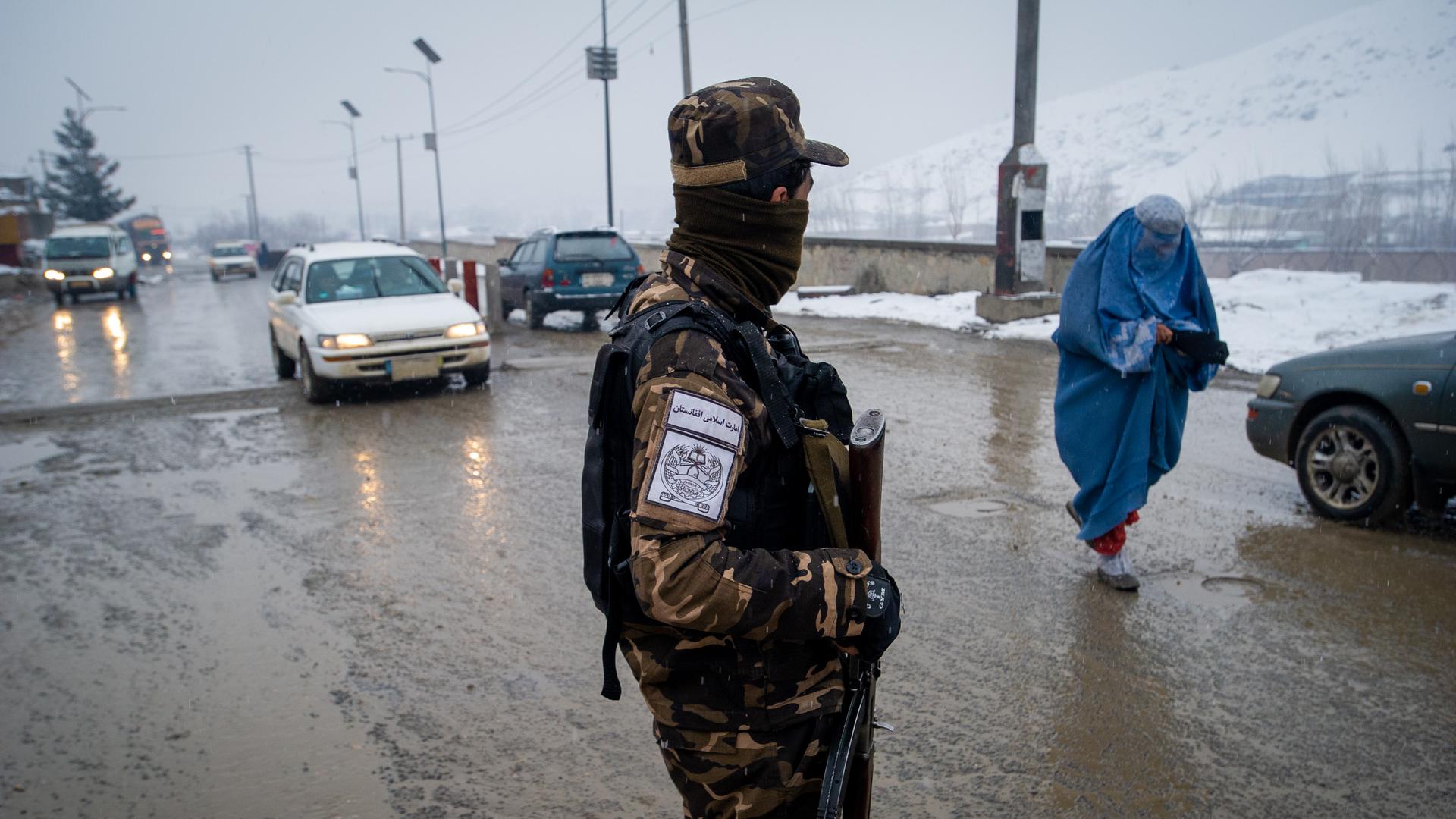 Eine afghanische Frau in blauer Burka bettelt im Schneegestäuber an einem Kontrollpunkt der Straße von Kabul nach Pul-e Alam, beobachtet von einem bewaffneten Taliban.