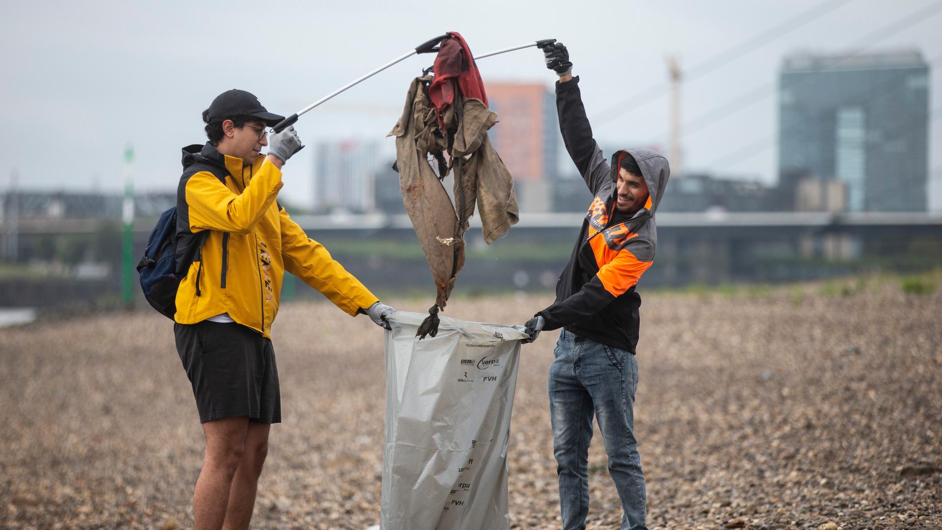 Zwei Männer stehen am Fluss-Ufer und tun Müll in eine Müll-Tüte. Zwei Männer stehen am Fluss-Ufer und tun Müll in eine Müll-Tüte.